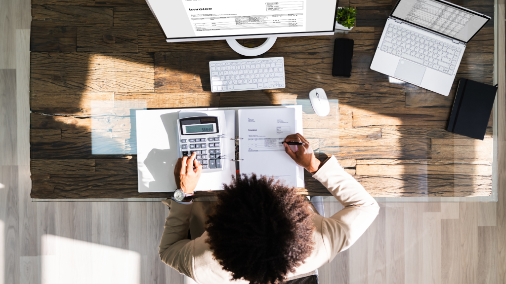 Accountant reviewing invoices and financial records using calculator and computer in modern office.
